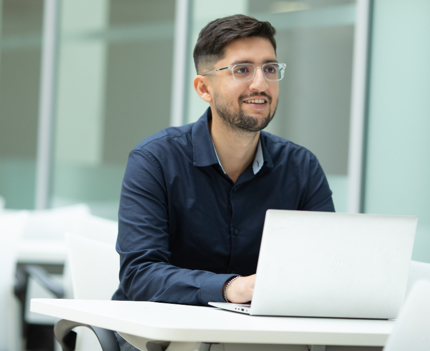 young man with glasses, working on a laptop and smiling just off camera 